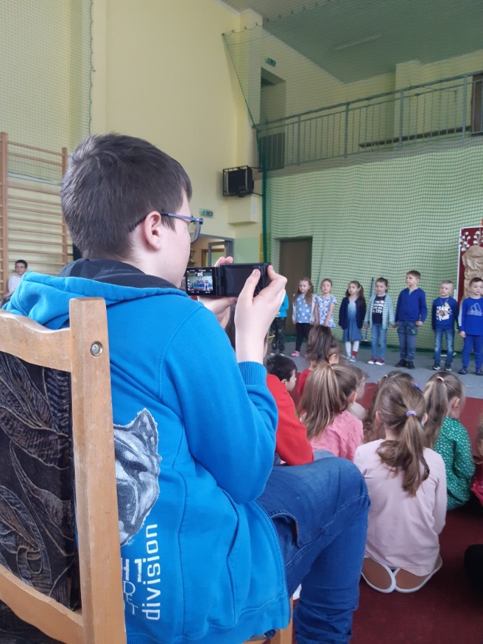 uczeń filmujący przedstawienie aparatem siedzący pośród innych uczniów szkoły