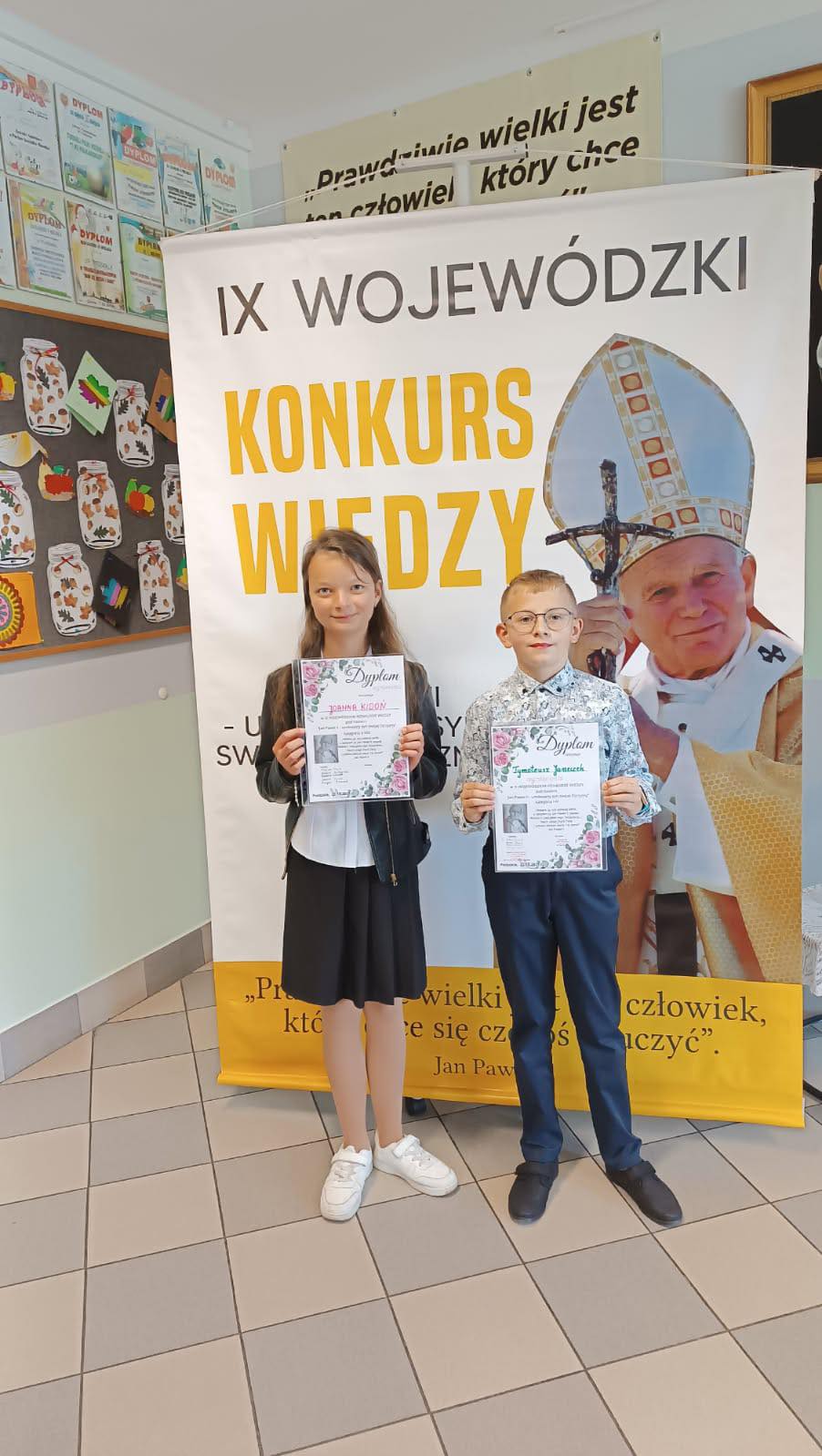 dwoje uczniów naszej szkoły z dyplomami na tle plakatu konkursu "Jan Paweł II - Umiłowany Syn Polskiej Ziemi"